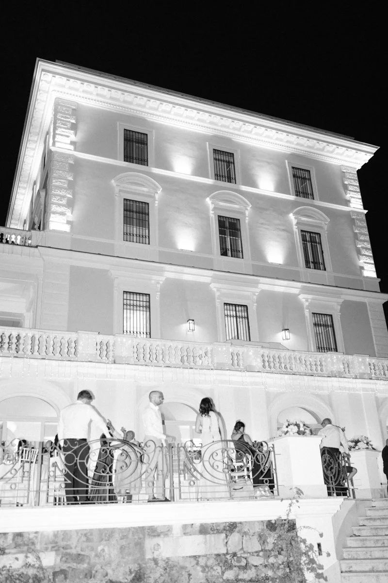 Nighttime photo of a large, three-story, classical-style building illuminated with exterior lights. Several people are gathered on a balcony or terrace in front of the building, some standing and some seated.
