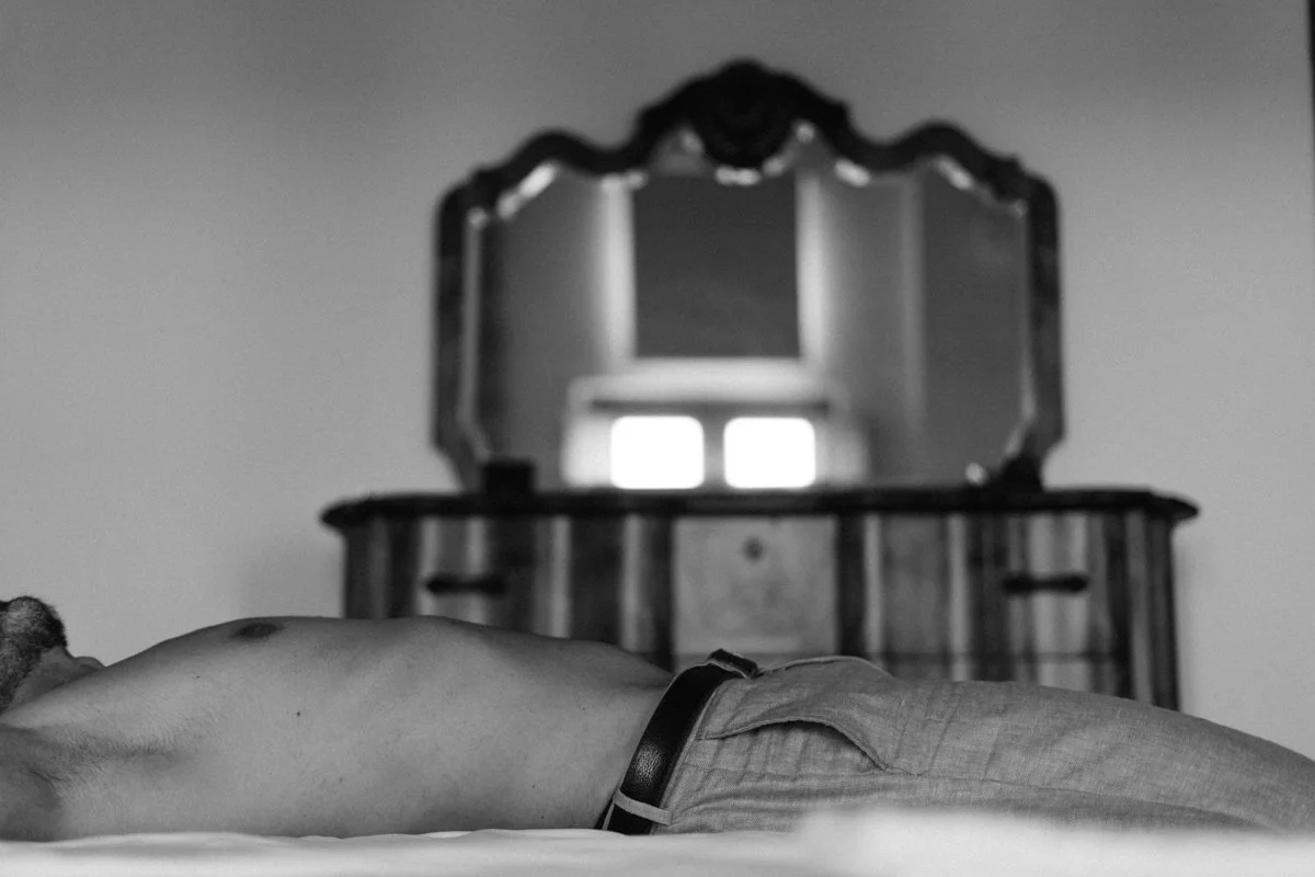 A scene with a person's bare back lying on a bed and a mirror reflecting a window in the background.