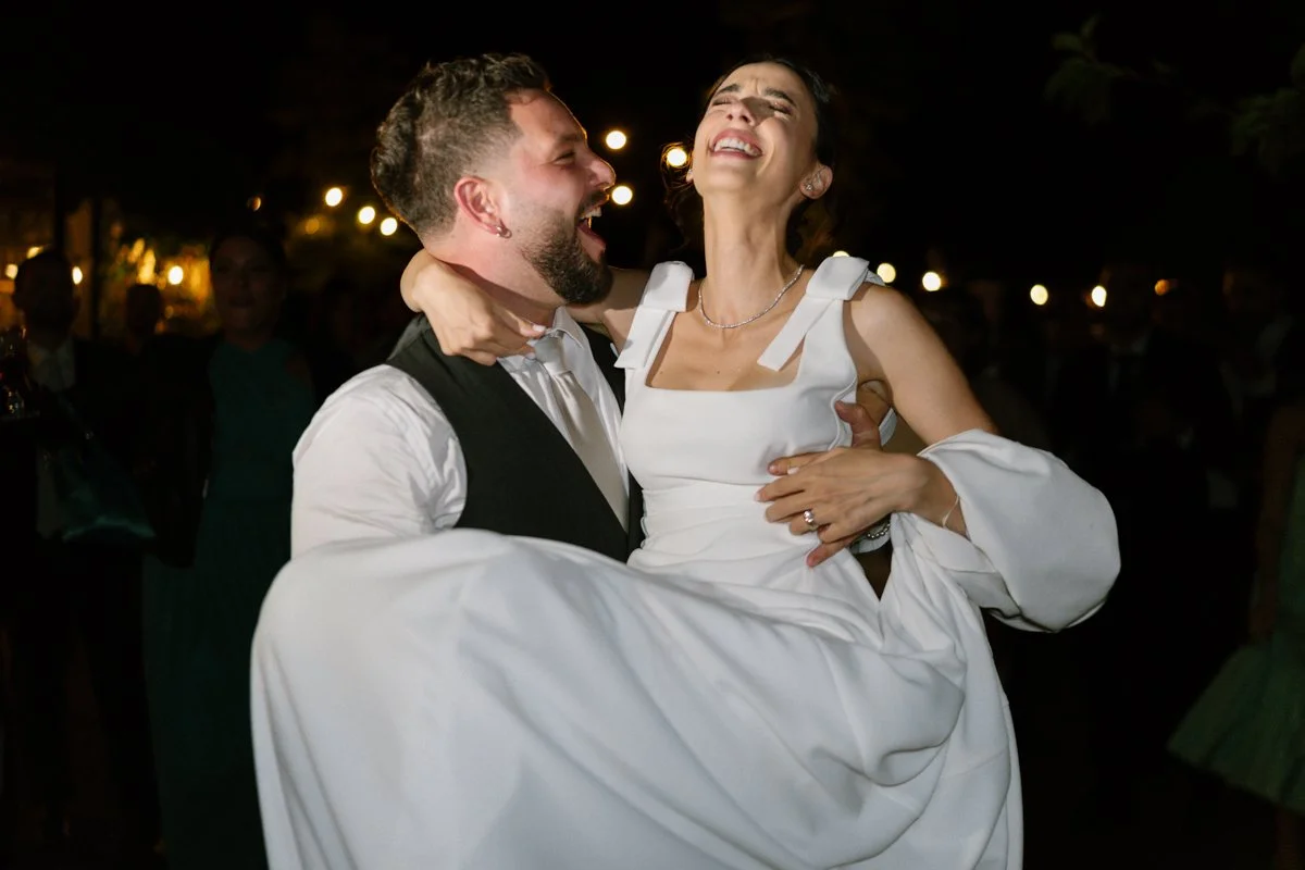 A man in a tuxedo lifts a woman in a white dress during a celebration at night, smiling and enjoying the moment.