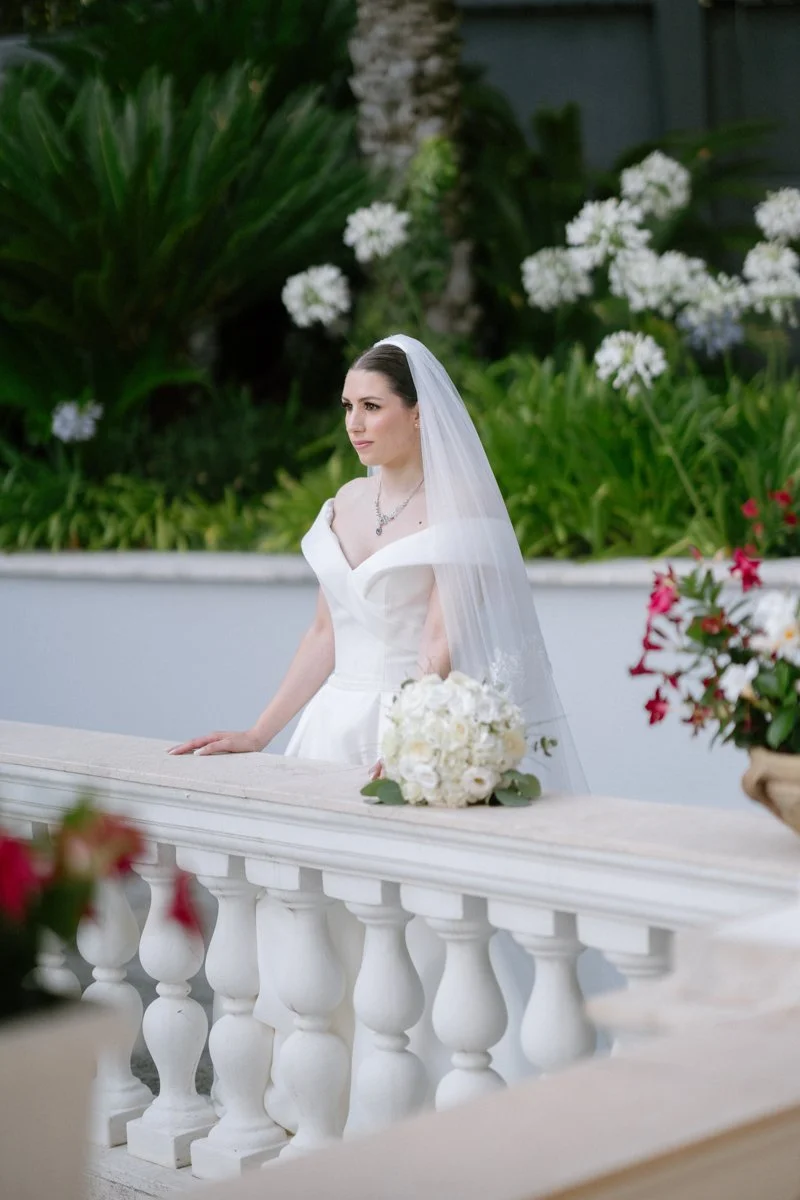 A bride in a white wedding dress with a veil standing outdoors next to a white balustrade, holding a bouquet of white flowers, with green foliage and white flowers in the background.