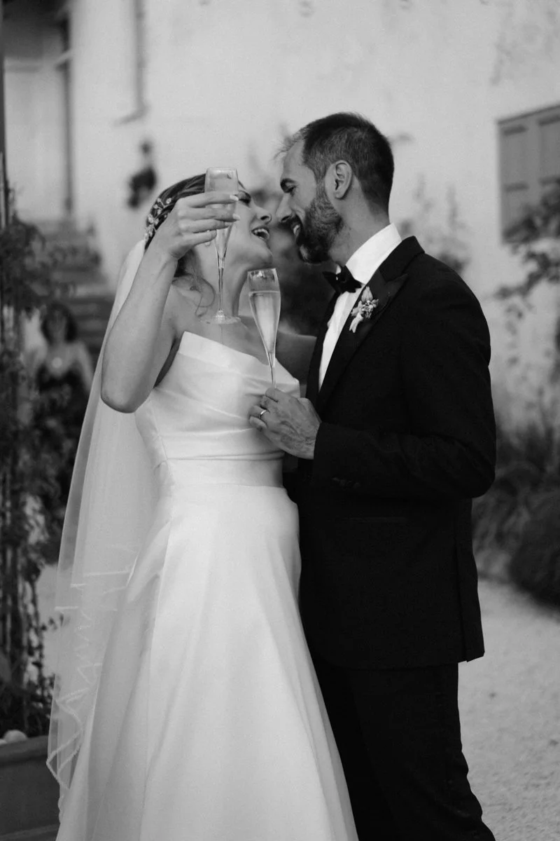 A bride and groom in wedding attire sharing a toast with champagne glasses, smiling and leaning close to each other.