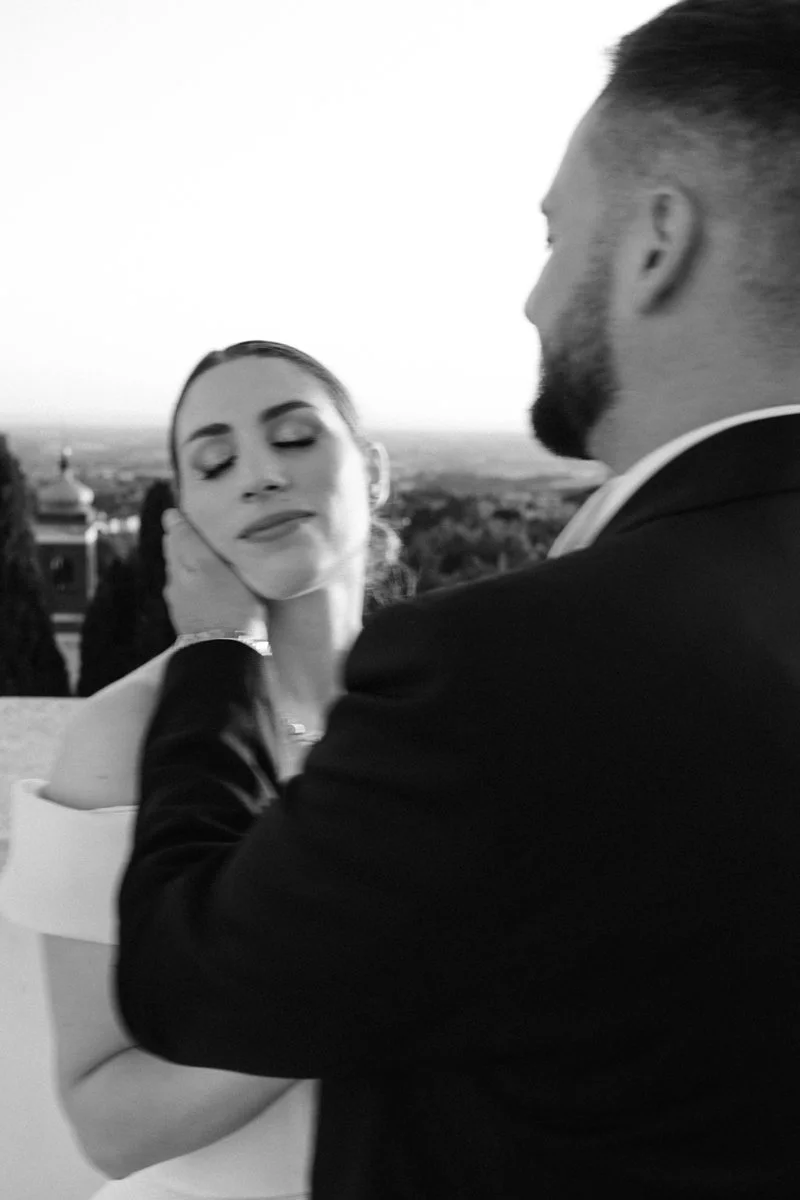 A black and white photo of a man in a suit holding a woman's face gently with both hands, her eyes are closed and she appears serene, with a scenic landscape in the background.