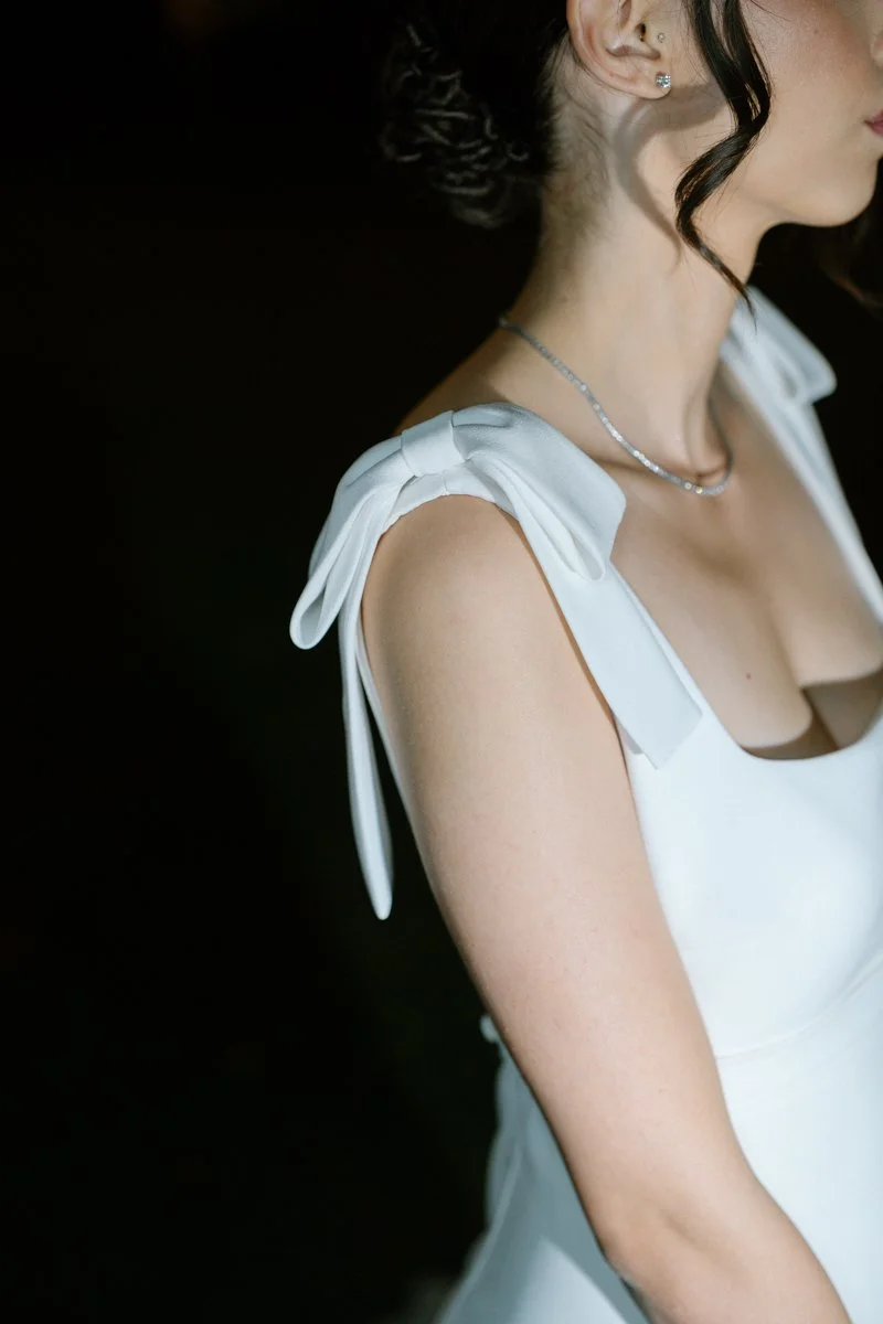 Close-up of a woman wearing a white dress with bow-tied shoulder straps, a silver necklace, and earrings, with her dark hair styled in an updo, against a dark background.