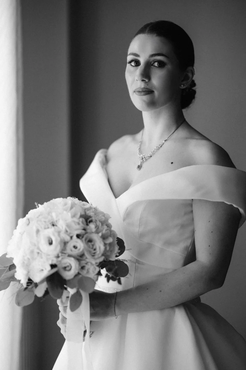 Black and white photo of a bride in a wedding dress holding a bouquet of flowers, looking at the camera.