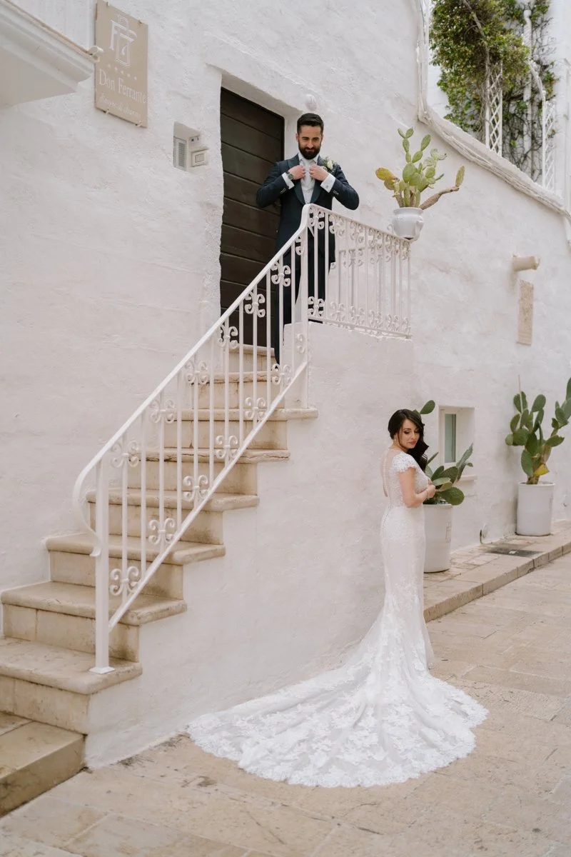 A bride in a white lace wedding dress with a train standing outside on a stone pathway, looking down, holding a green plant, near a white wall with potted cactus plants. A groom in a dark suit is on the stairs above her, adjusting his bow tie.