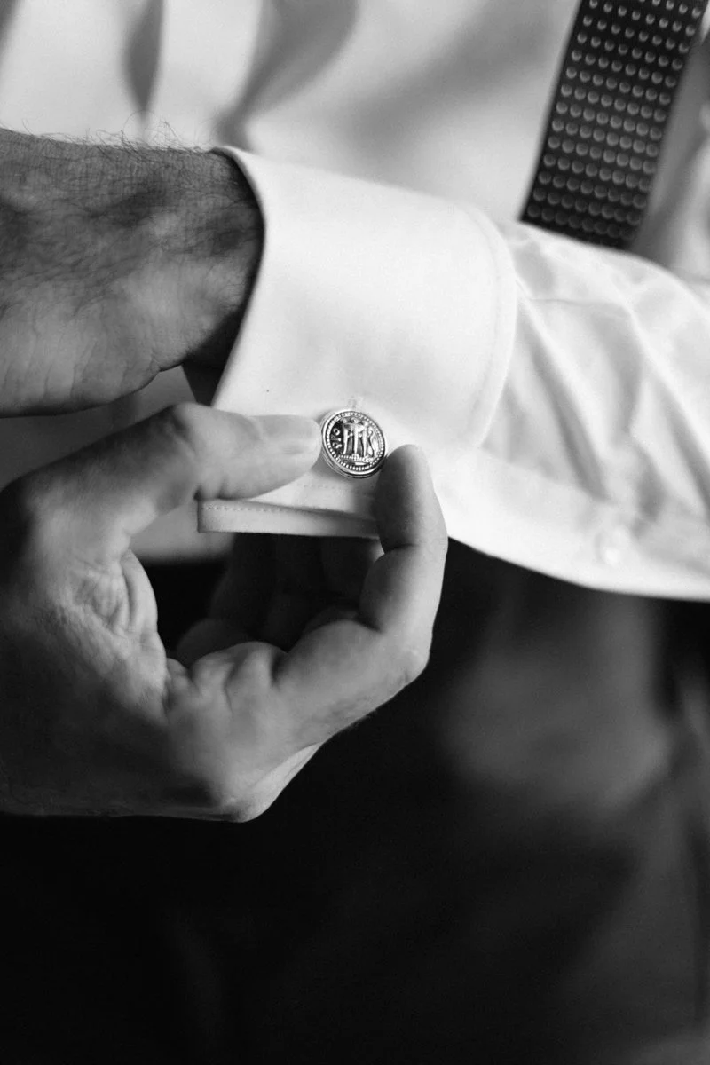 Close-up of a man's hand adjusting a cufflink on a white dress shirt, with a patterned tie visible in the background, in black and white.