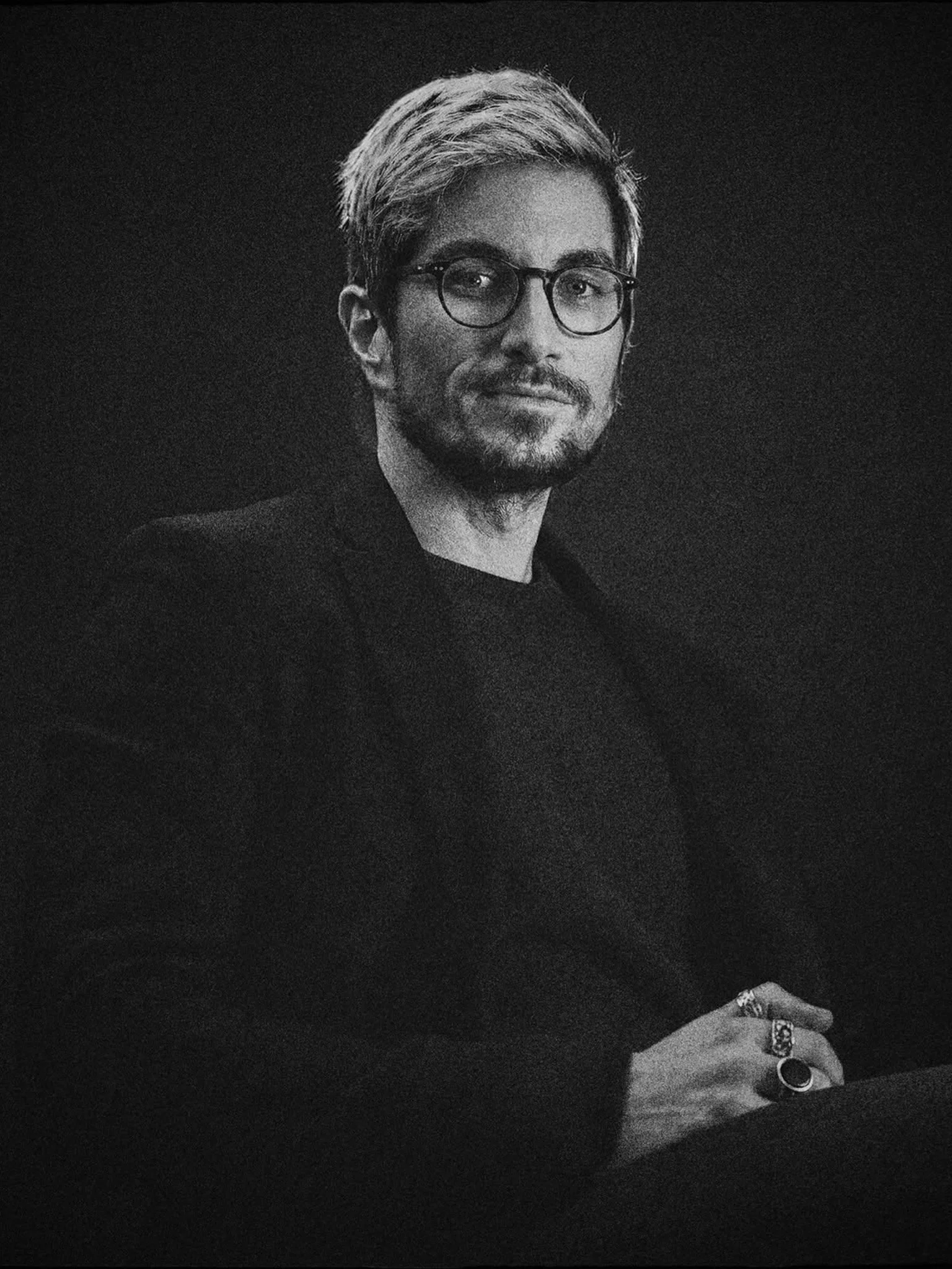 Black and white photograph of a man with a beard, glasses, and styled hair, wearing rings and a dark outfit, resting his hand with rings on a surface against a dark background.