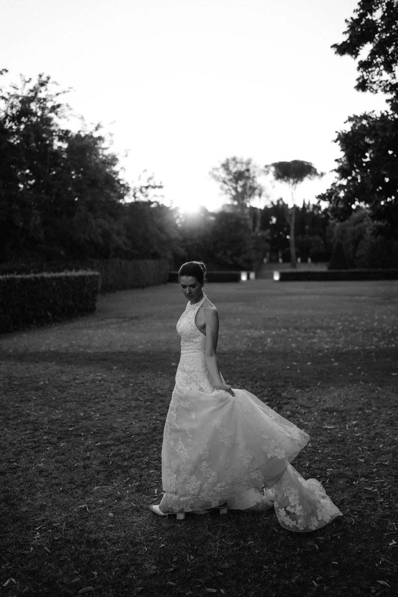 A woman in a lace wedding dress standing on a grassy field at sunset, holding the edge of her dress and looking down, with trees and a park in the background.