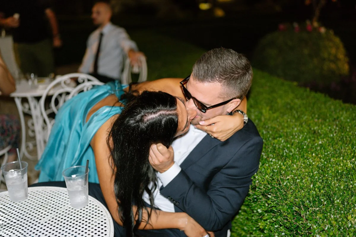 A man in a suit and glasses and a woman in a blue dress sharing a kiss at a nighttime outdoor event, with drinks on a nearby table and other guests in the background.