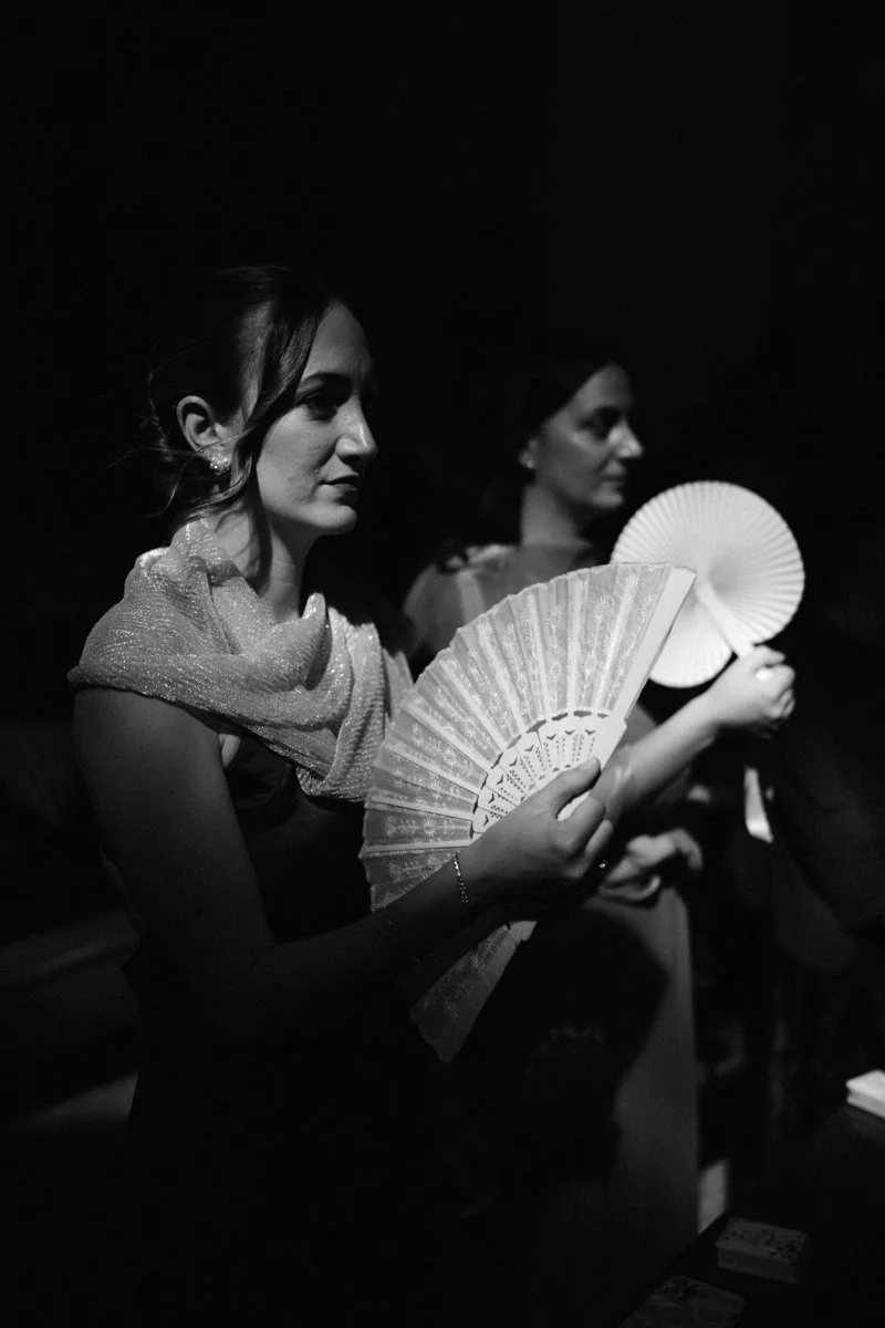 Two women holding paper fans in a dark setting, with the foreground woman in focus and the background woman slightly blurred, both with dark hair and elegant attire.