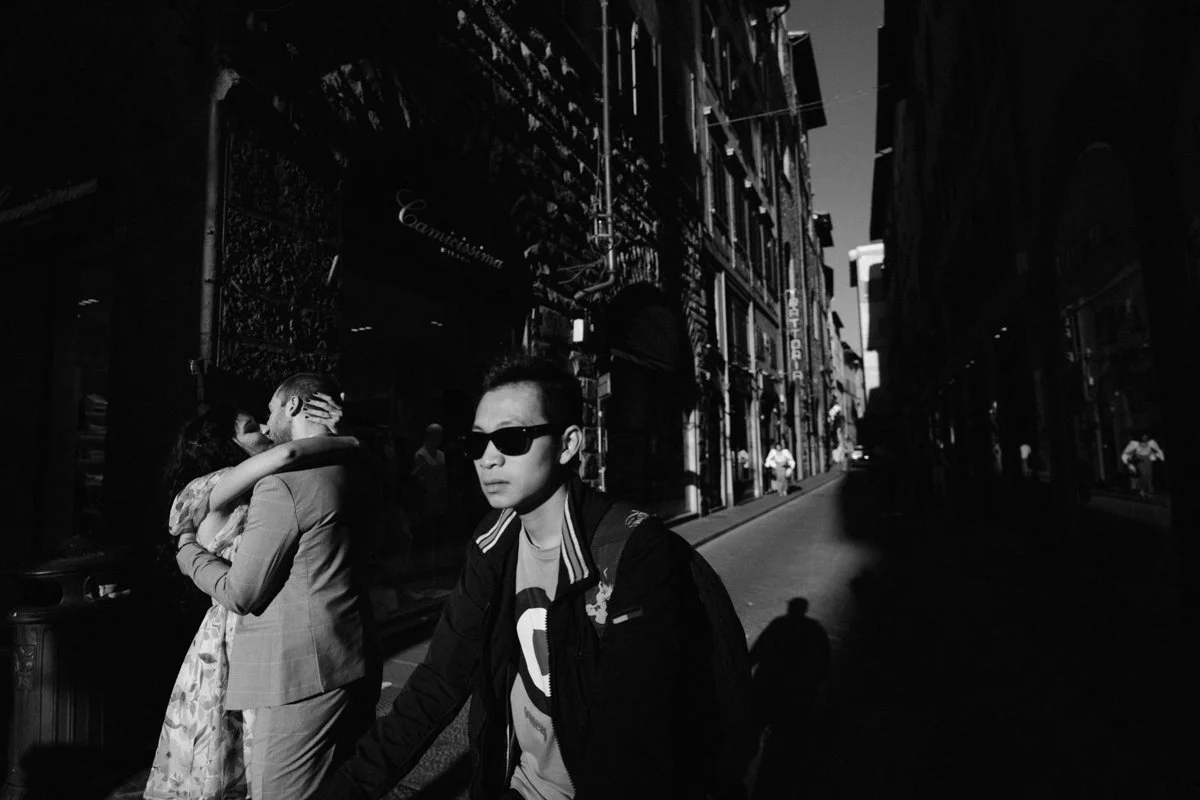 A man with sunglasses and a backpack walking on a city street, with a couple embracing in the background and a few pedestrians. The scene is in black and white.