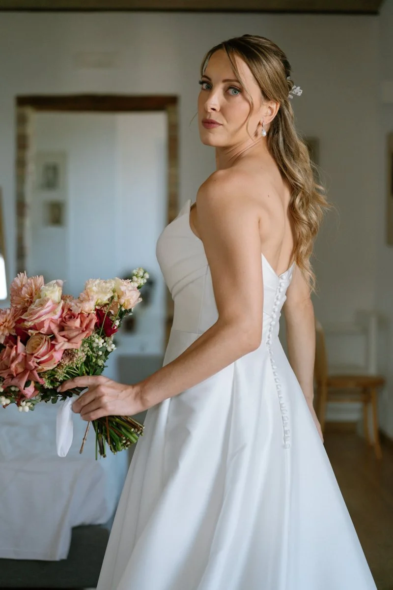 A bride in a white strapless wedding dress holding a bouquet of pink and white flowers, standing indoors with a background of white walls and framed pictures.
