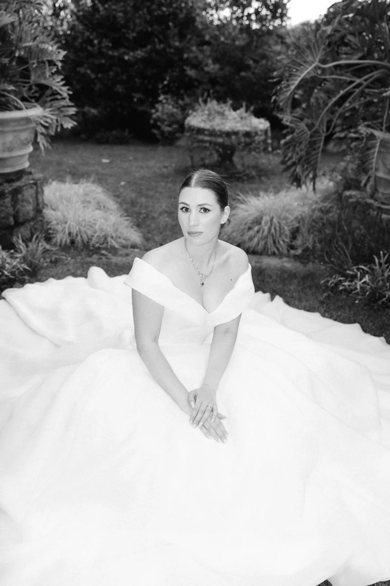 A woman in a white off-shoulder gown sitting outdoors in a garden setting with plants and trees in the background.