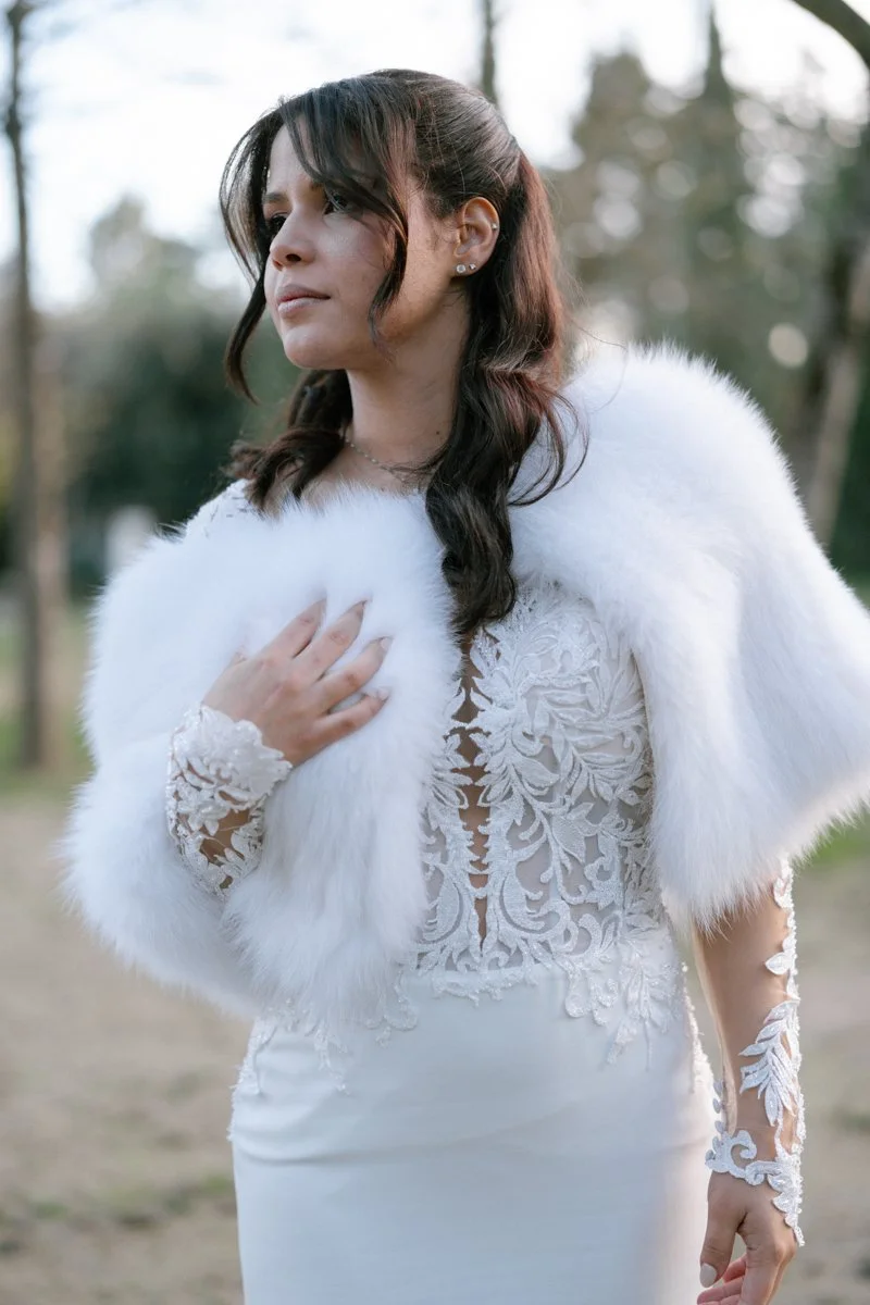Woman in a white lace dress with a furry white shawl, standing outdoors with blurry trees in the background.
