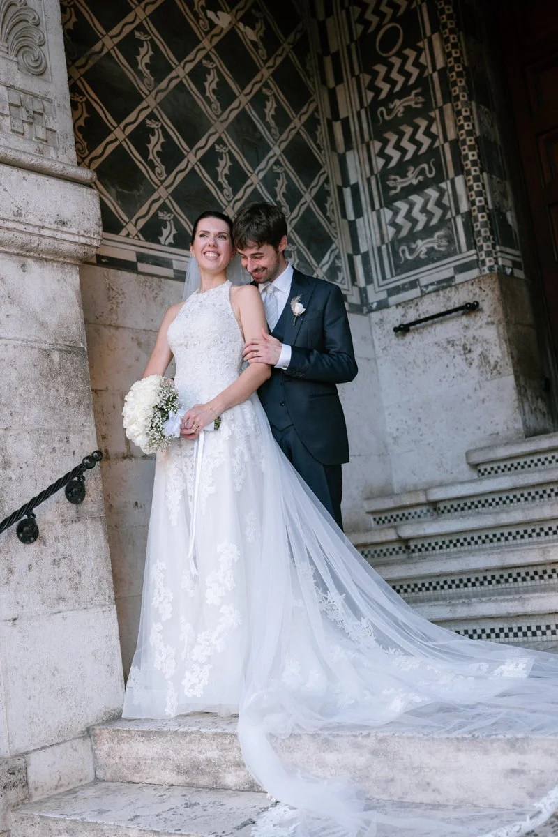 A bride and groom standing on the steps of a historic building outside, smiling and holding each other, with the bride holding a bouquet of white flowers, during their wedding celebration.