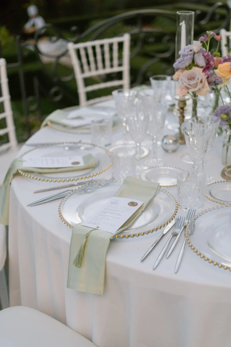 Formal outdoor table setting with white tablecloth, clear glass plates with gold beaded rims, crystal glassware, silver cutlery, and a floral centerpiece with pastel roses and other flowers. Napkins have menus, and chairs with white covers are around