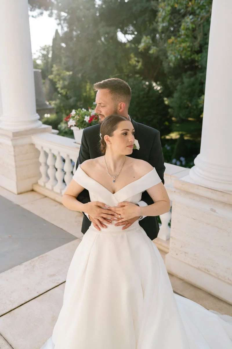 A newlywed couple standing on a balcony with white columns, surrounded by lush greenery. The bride is in a white off-shoulder wedding gown with jewelry, and the groom is in a black suit with a white shirt, embracing her from behind.