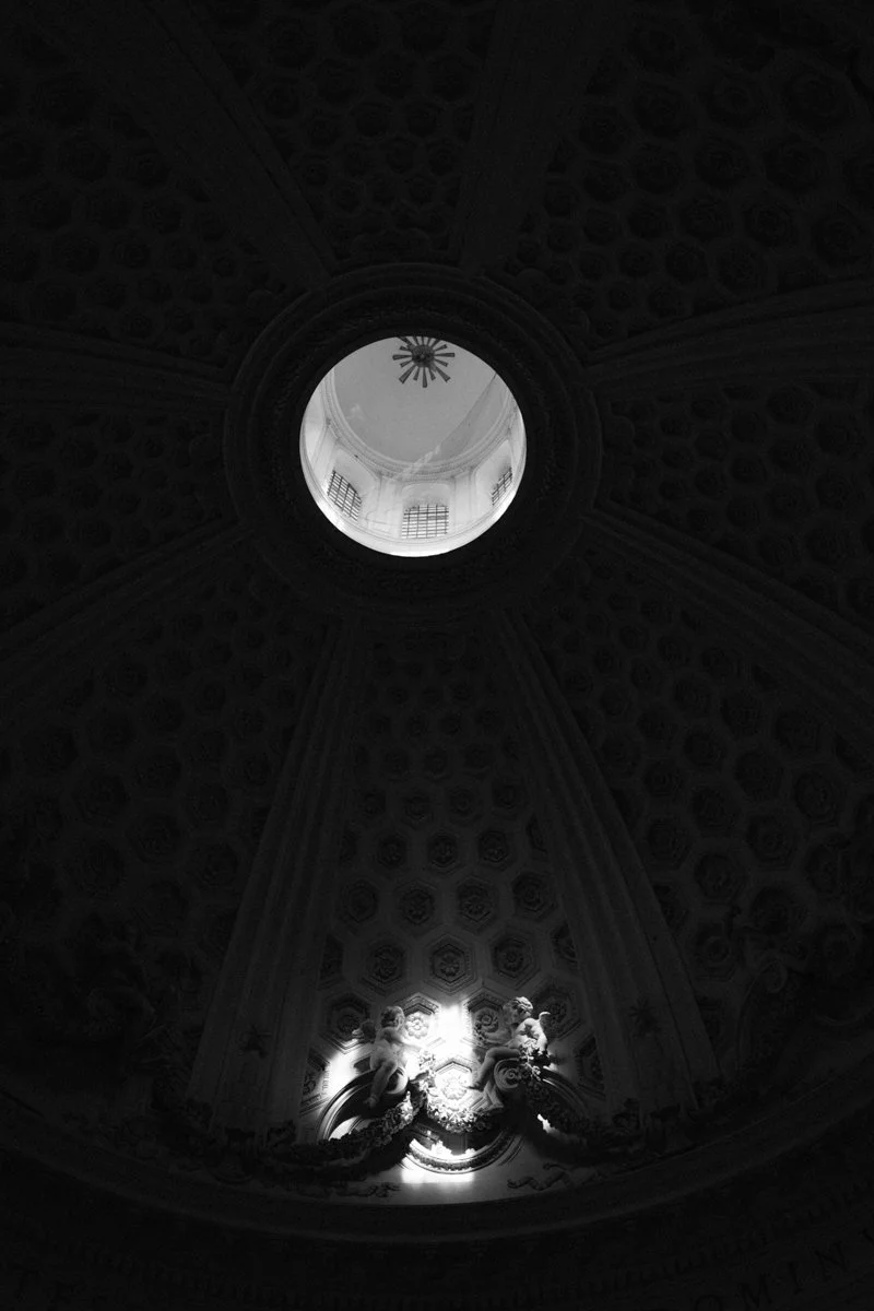 Black and white photo of the ceiling inside a dome with an oculus at the center, showcasing intricate architectural details and statues illuminated by light.