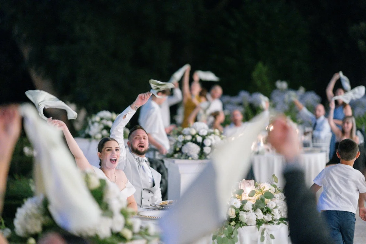 People celebrate at a wedding reception outdoor at night, with guests waving napkins in the air, surrounded by floral arrangements and tables.