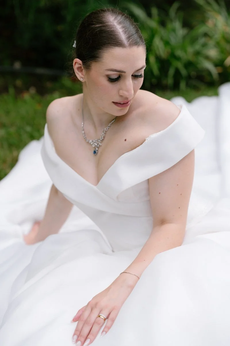 A bride in a white wedding dress sitting outdoors, looking down, with a lush green background.