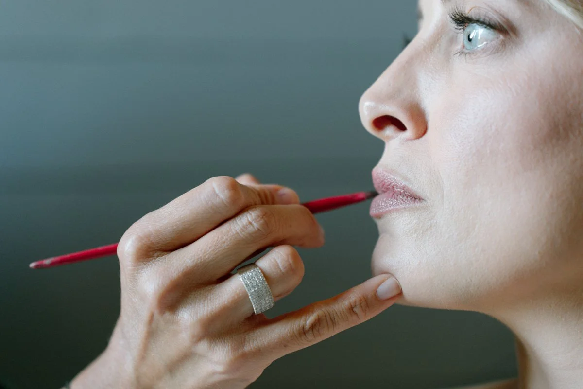 Close-up of a woman with blue eyes having makeup applied to her lips with a red brush, held by a makeup artist's hand wearing a silver ring.
