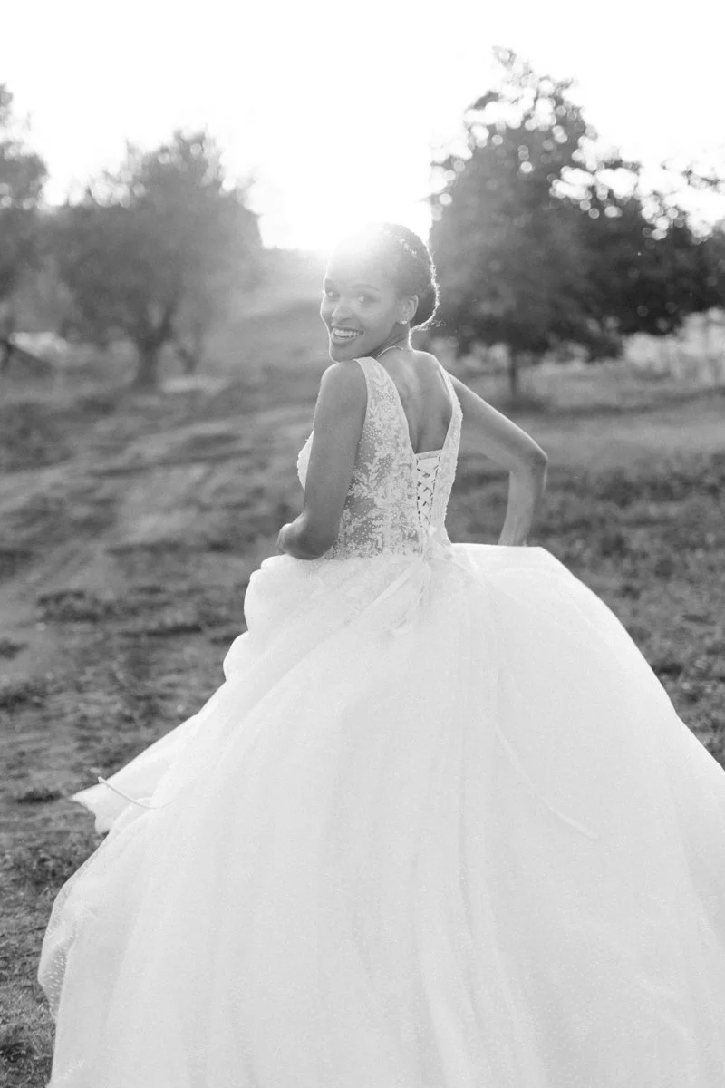 A smiling woman in a wedding gown walking outdoors during sunset with trees in the background.