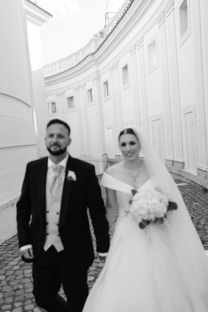 A black-and-white photo of a bride and groom walking hand in hand outdoors near a white curved building, the bride holding a bouquet and wearing a wedding dress, and the groom in a tuxedo.