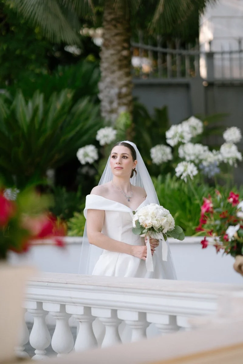 A bride with dark hair in a white off-shoulder wedding gown holding a white bouquet, standing outdoors with green plants and white flowers in the background.