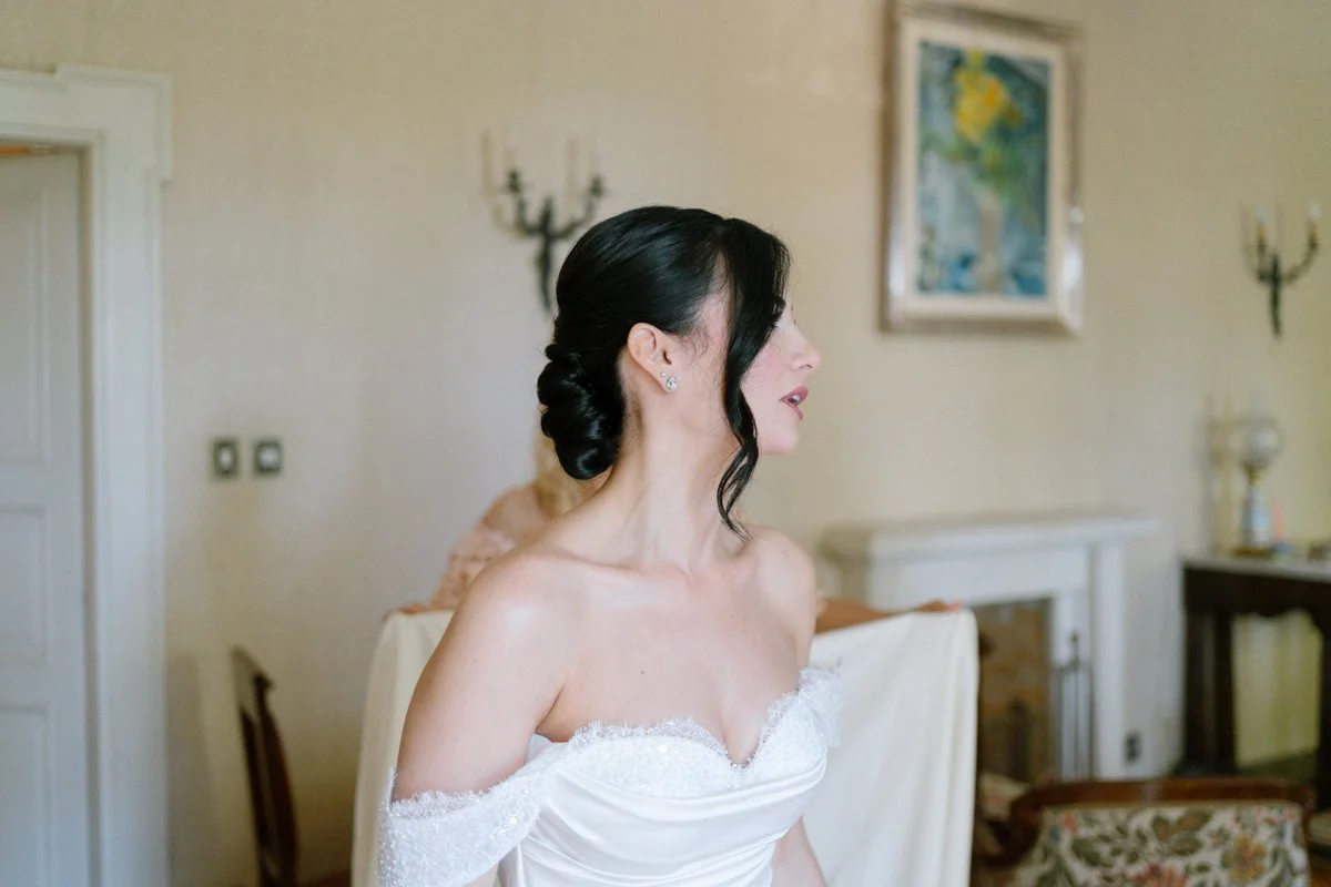 A woman with dark hair in an elegant updo, wearing a white off-shoulder dress, sitting in a room with beige walls, a framed painting, and vintage-style furniture.