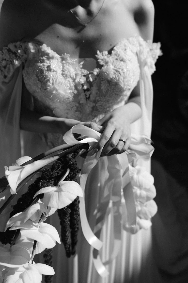 Black and white photo of a bride holding a bouquet of white flowers, wearing a strapless wedding dress with lace detailing.