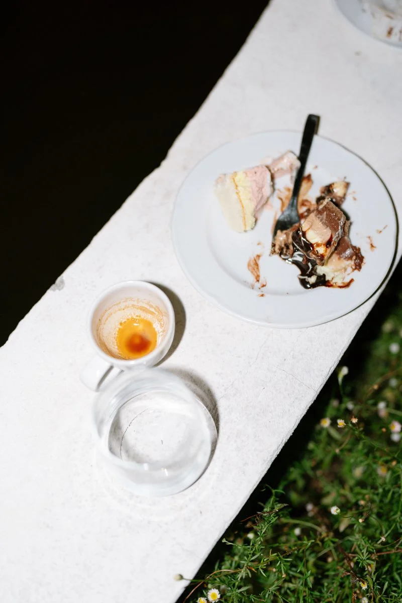 A white plate with leftover slices of cake, a spoon, a small cup with coffee or espresso, and an empty glass on a white surface outdoors.