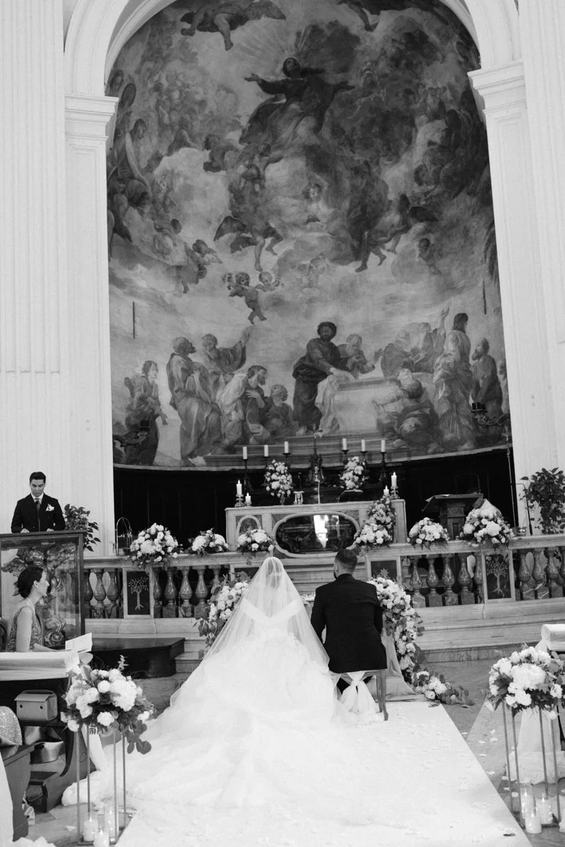 A bride and groom kneeling at the altar during a wedding ceremony inside a church with a large religious mural painting on the wall behind them, decorated with flowers and candles.