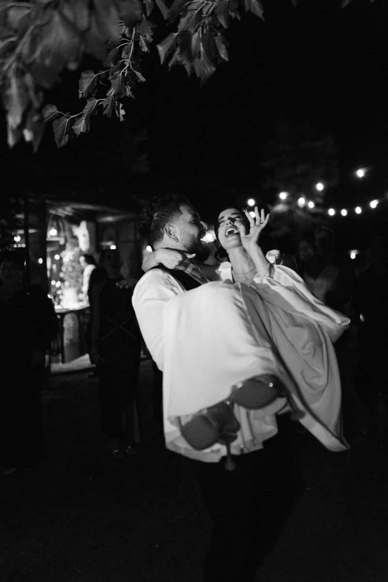 A man is holding a woman in his arms at an outdoor night event with string lights, and they are laughing and enjoying themselves.