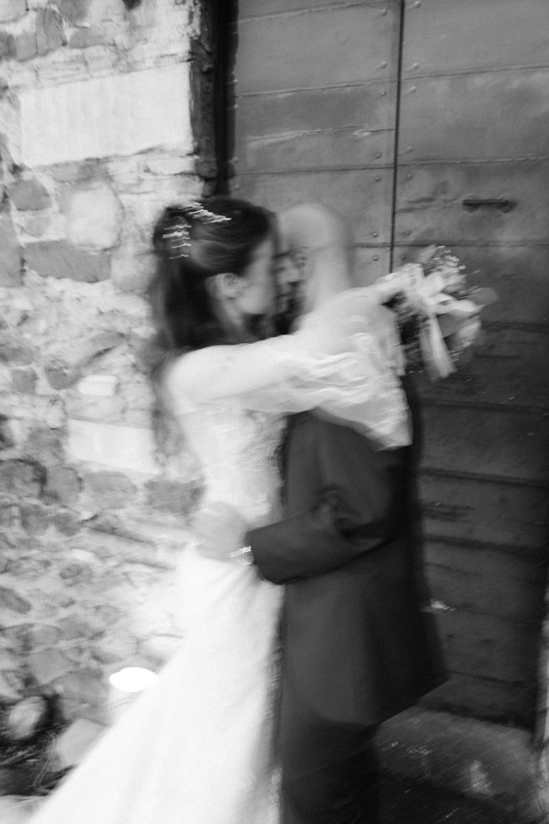 A romantic couple, wearing wedding attire, embracing and sharing a kiss indoors with a rustic brick and wood wall background.