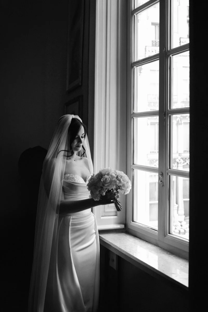 A bride in a strapless wedding dress and veil holding a bouquet of flowers, standing by a window with natural light.