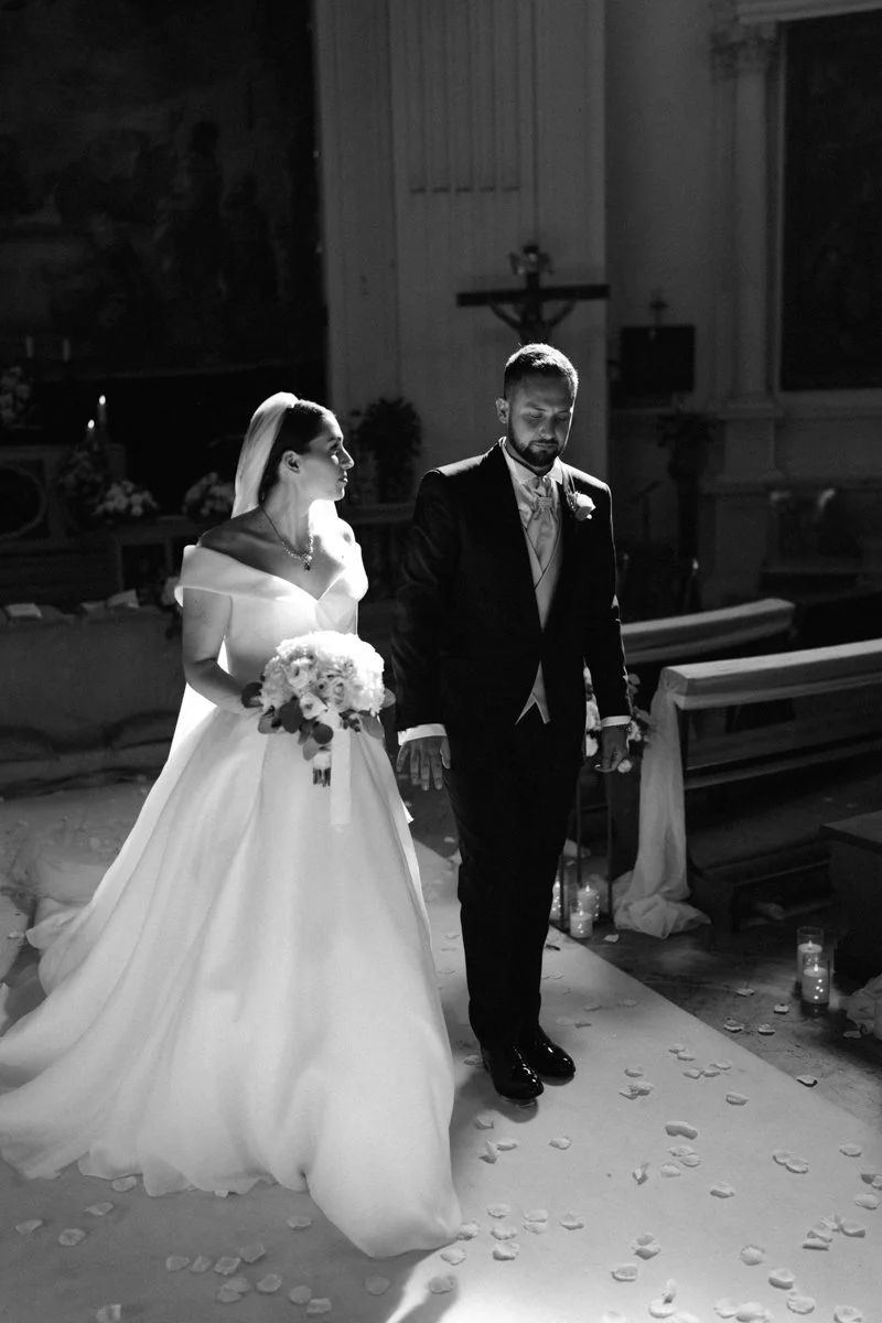 Black and white photo of a bride and groom standing at the altar during a wedding ceremony inside a church. The bride is holding a bouquet of flowers, wearing a wedding gown and veil, while the groom is dressed in a tuxedo. There are candles and flow