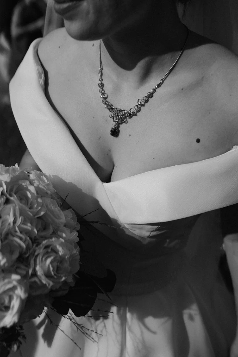 Close-up of a woman wearing a necklace and a strapless dress, holding a bouquet of flowers, with a focus on her chest and face in black and white.