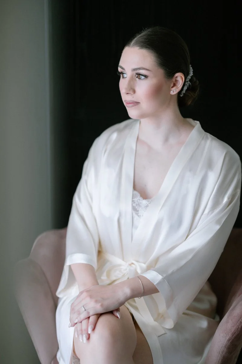 A woman with her hair styled in an elegant bun, wearing pearl earrings, and a silky, cream-colored robe over lace lingerie, sitting on a chair near a window, gazing thoughtfully outside.