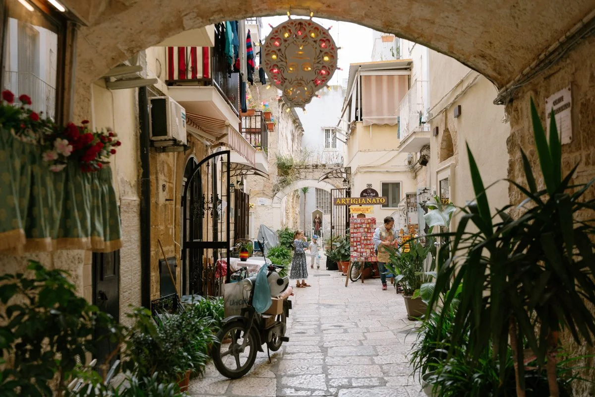 View of a narrow European street through an archway, with shops, potted plants, people chatting, and a sign reading 'Artigianato'.