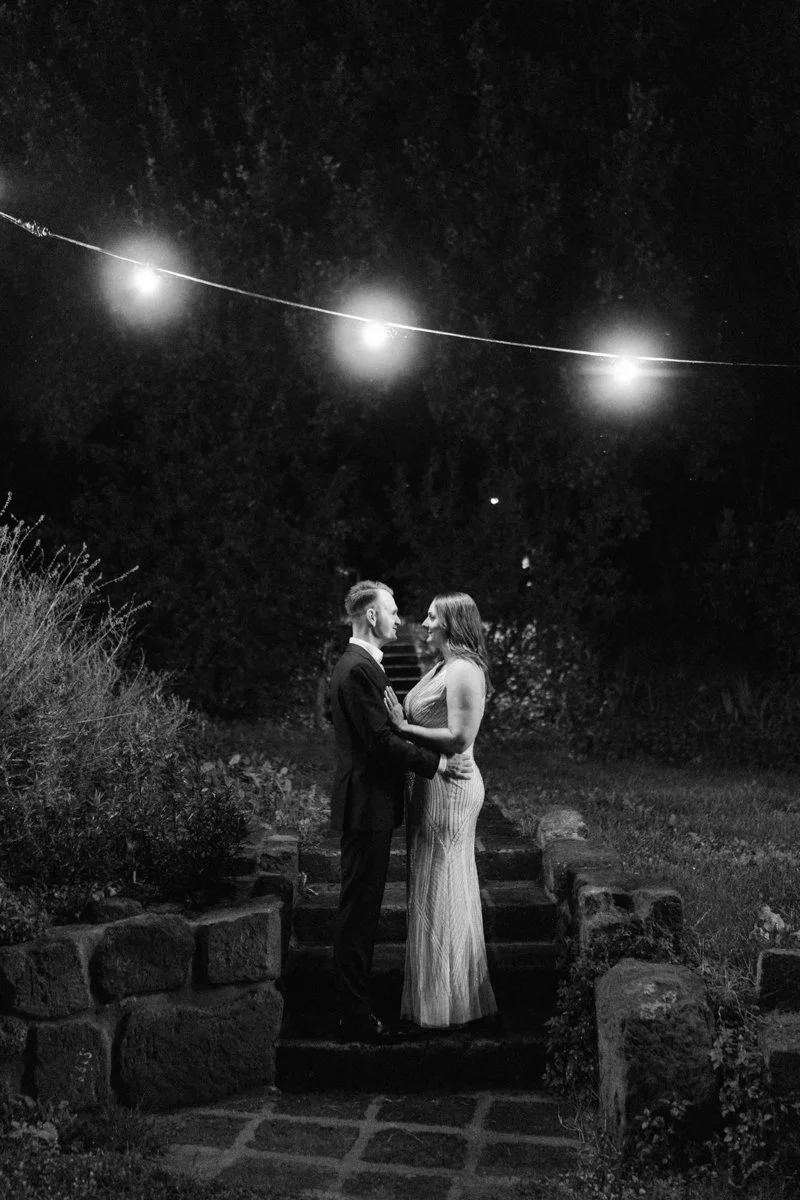 A black and white photo of a couple standing on steps outdoors at night, gazing into each other's eyes under string lights hanging overhead.