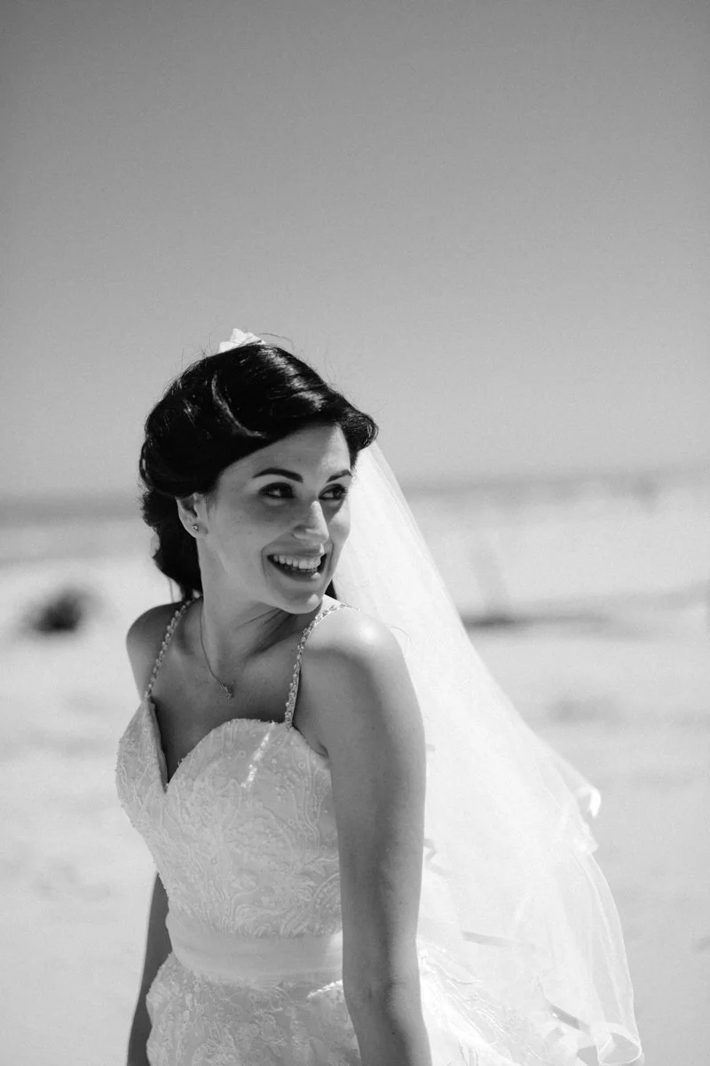 Black and white photo of a smiling bride on a beach, wearing a wedding dress and veil.