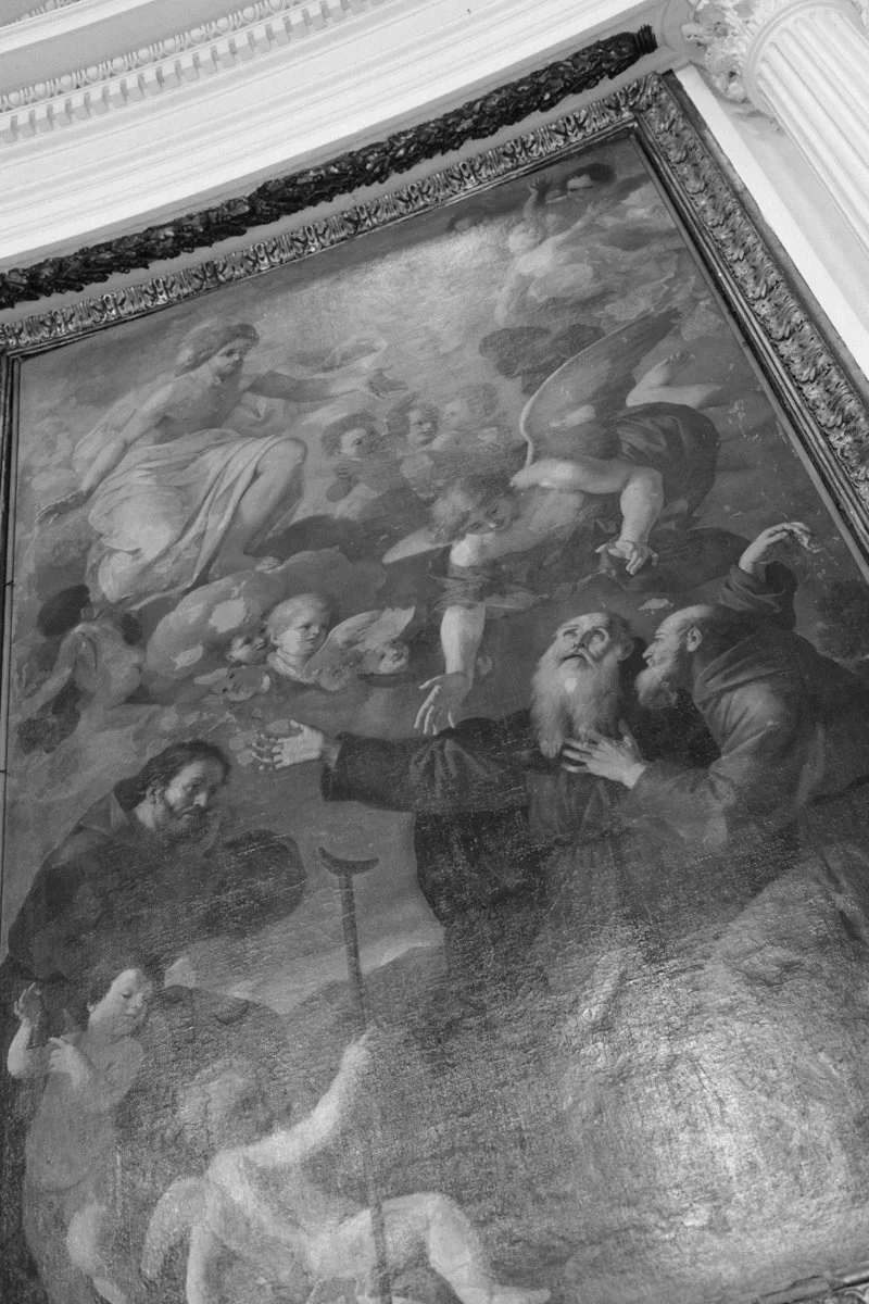 A framed classical painting or artwork depicting several mythological or historical figures, including winged angels or gods, in a dynamic scene with some figures reaching out and interacting, set against a detailed ornate ceiling background.