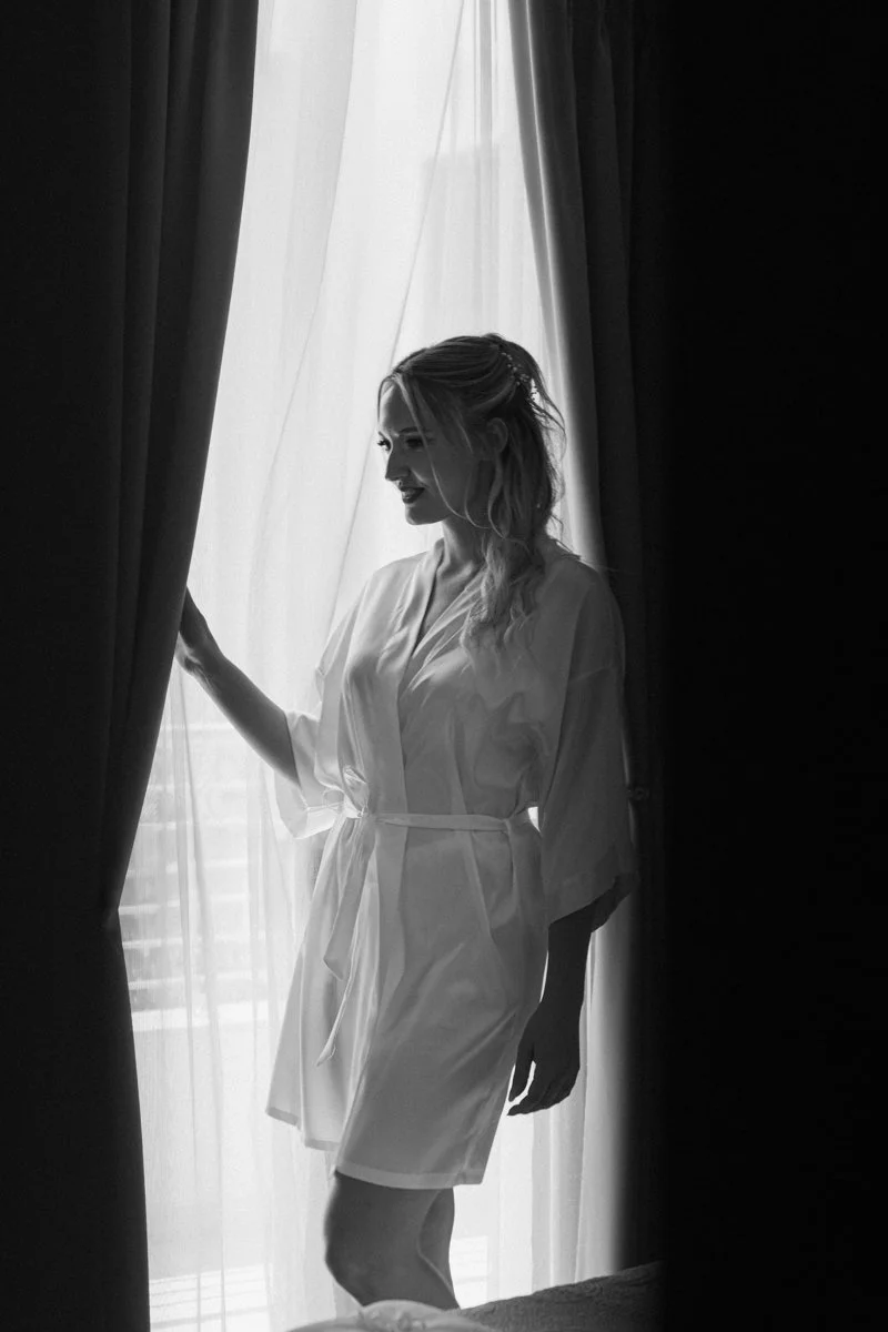 A woman in a robe standing by a window, holding back a curtain and smiling, in black and white.