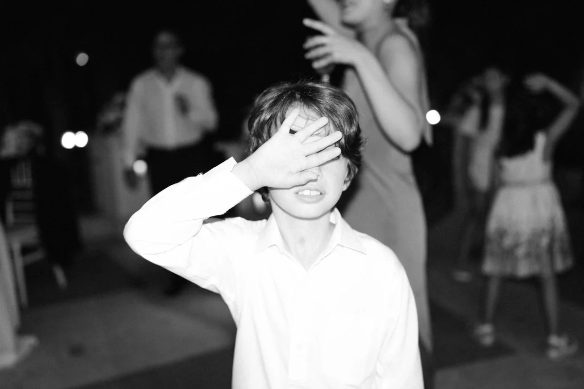 A boy is covering his eyes with his hand at a party or gathering, with people in the background.
