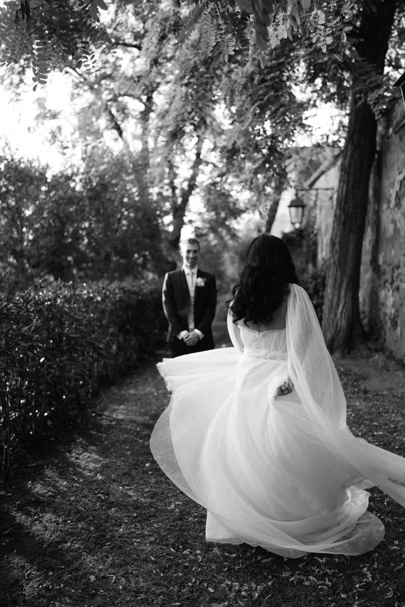 A black-and-white photo of a bride in a flowing wedding dress running towards a smiling groom, outdoors over a dirt path lined with bushes and trees.
