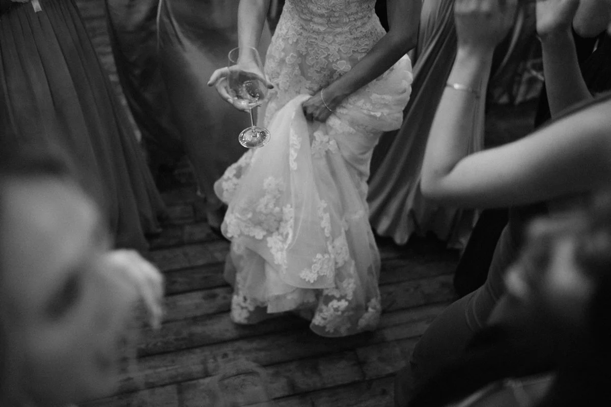 A bride holding a glass of wine lifts her wedding dress while dancing at her wedding reception, surrounded by guests.