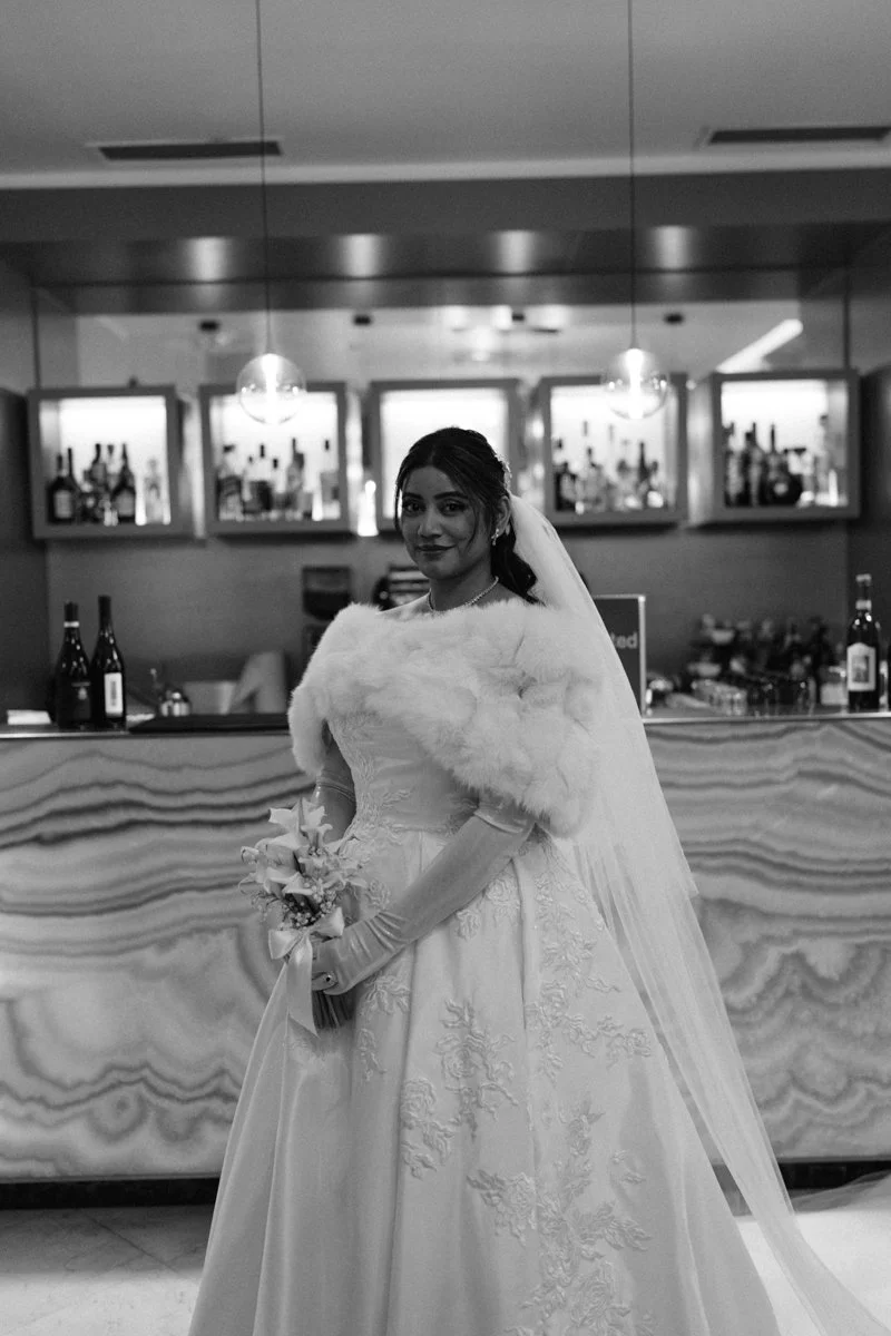 A bride in a wedding dress holding a bouquet, standing indoors in front of a bar with bottles on display behind her.