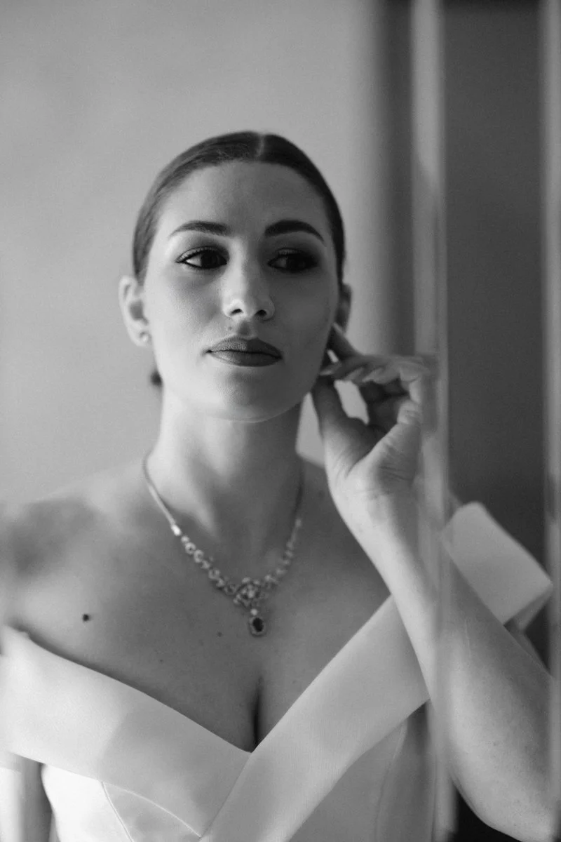 Black and white portrait of a woman with styled hair, wearing a necklace and an elegant off-shoulder dress, standing near a mirror and touching her ear.