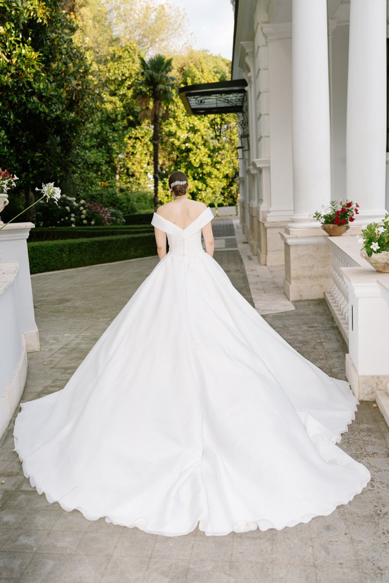 A bride in a white wedding gown with an off-the-shoulder design and a long train walking outdoors on a paved pathway, with greenery and white columns in the background.
