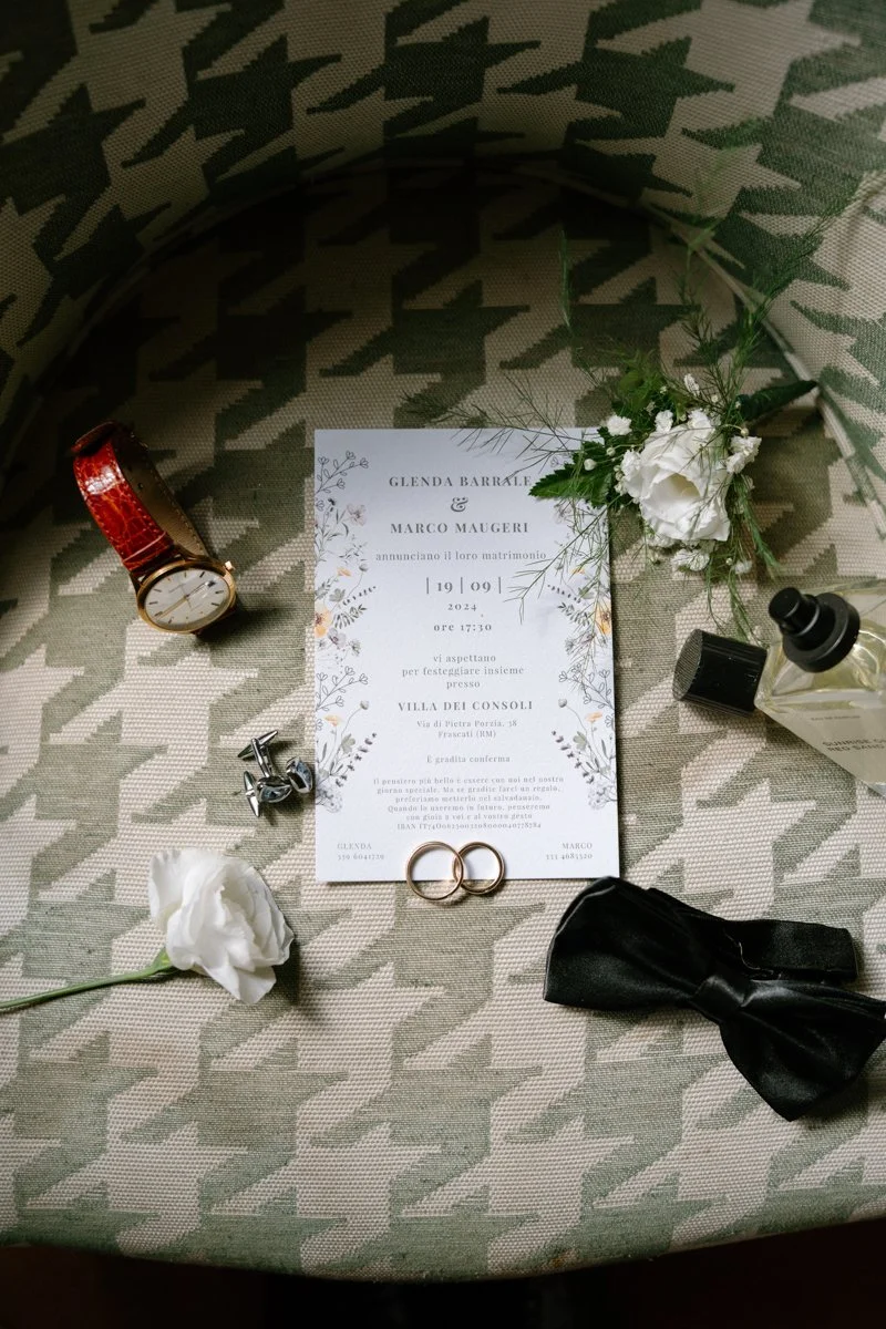 Wedding invitation surrounded by a wristwatch with a red strap, a pair of wedding rings, a white flower, a black bow tie, a boutonniere, and a bottle of perfume on a patterned fabric surface.